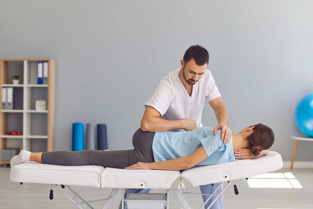 Clinician assessing a person’s back posture while the person stands in a bright examination room.